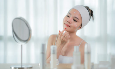 Portrait d'une belle jeune femme asiatique prenant soin de sa peau assise près du miroir dans la chambre avec une bouteille de soins de la peau sur la table, femme touchant le visage.